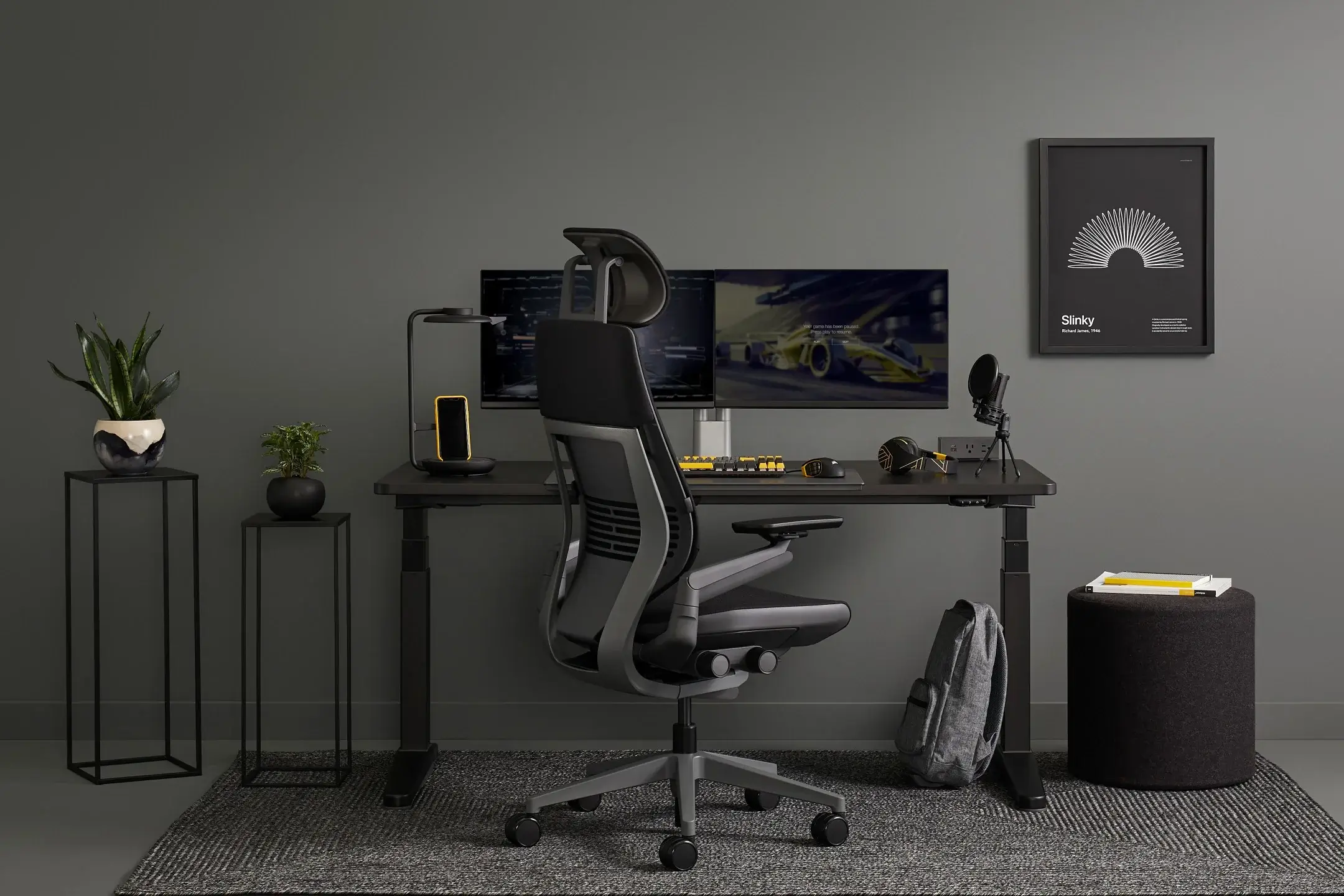 Ergonomic home office with a black desk, dual monitors, a microphone, and a framed poster on a gray wall.